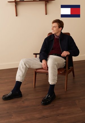 Een jonge man zit op een houten stoel, gekleed in een marineblauw bomberjack, een bordeauxrode trui, grijze joggers, zwarte loafers en marineblauwe sokken.