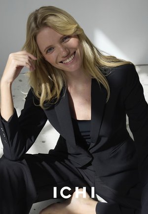 Black tailored blazer with padded shoulders, matching trousers, worn over a black top. Smooth fabric, fitted silhouette, and minimalistic design.