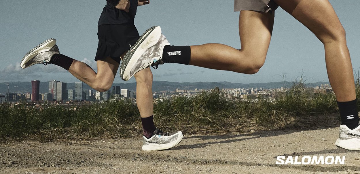 Due corridori con scarpe Salomon corrono a tutta velocità su un sentiero di terra con sullo sfondo lo skyline della città e le montagne.
