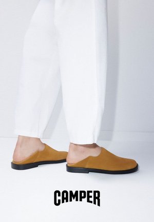 Persona che indossa pantaloni bianchi e scarpe slip-on color cuoio, in piedi su una superficie bianca con il logo del marchio Camper sotto.