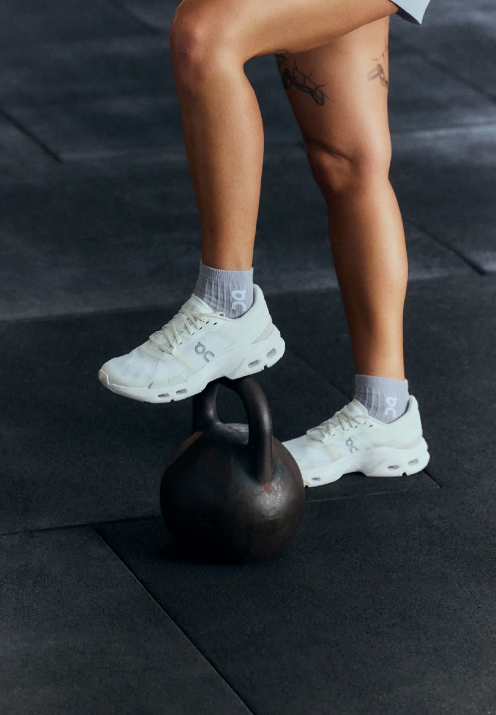 Weiße Sportschuhe mit durchlöcherten Einsätzen und einer gepolsterten Sohle auf einem schwarzen Kettlebell, mit grauen Socken mit Logo, auf dunklem Boden im Fitnessstudio.