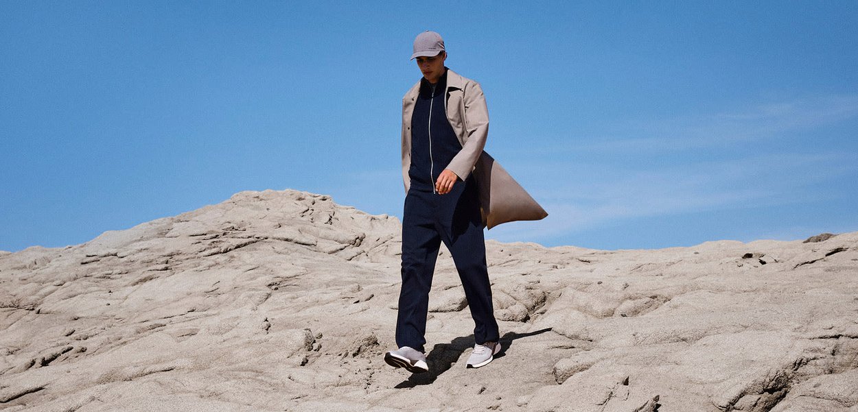 Uomo che indossa un cappotto beige, abbigliamento blu navy e un berretto mentre cammina su un terreno roccioso sotto un cielo azzurro limpido.