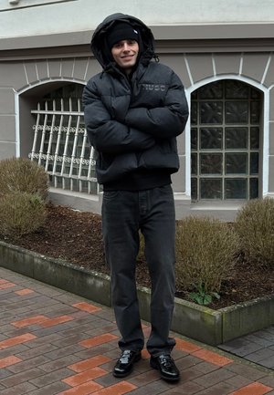 Giacca imbottita nera con cappuccio, logo sul petto, abbinata a jeans neri e scarpe lucide. In piedi sul marciapiede con piante e uno sfondo di edificio.