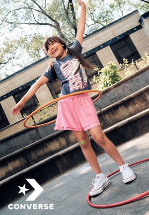 El conjunto para niños de Converse incluye una camiseta gris con un gráfico de zapatillas, pantalones cortos de deporte rosa y zapatillas de lona blancas, en acción con un hula hoop.