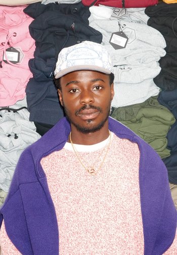 Casual outfit met een roze gespikkelde trui, een paarse buitenlaag en een geblokt petje. Achtergrond van keurig gestapelde hoodies in verschillende kleuren.