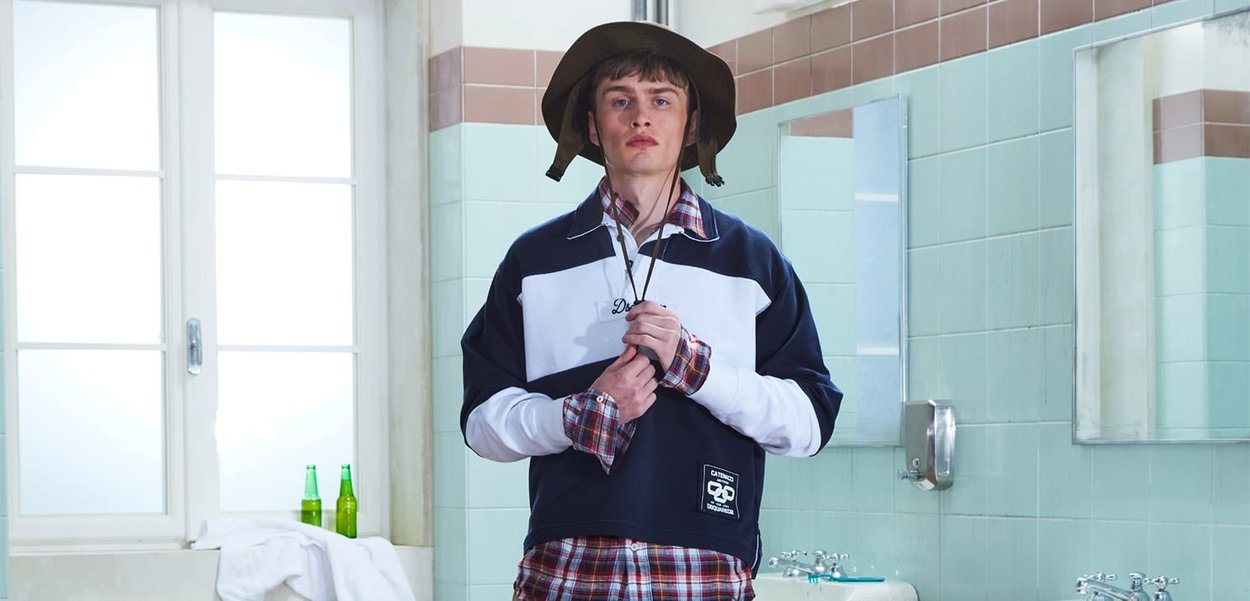 Joven con sombrero de ala ancha y ropa casual en capas, de pie en un baño con azulejos, sosteniendo el cuello de su chaqueta cerca del lavabo y el espejo.