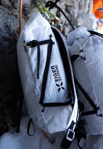 Weiße Eiger Extreme Kletterrucksäcke mit schwarzen Trägern, die an einer felsigen Wand im Schnee lehnen.