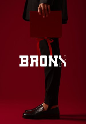 Schwarze Leder-Halbschuhe mit niedrigem Profil, kombiniert mit maßgeschneiderten Hosen. Eine rote Geschenkbox mit Schleife wird in einer Hand vor einem dunkelroten Hintergrund gehalten.