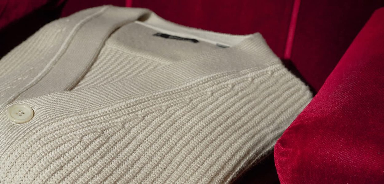 Cardigan color crema con un design a coste testurizzato, caratterizzato da scollo a V e un bottone rotondo bianco, posato su una superficie di velluto rosso.