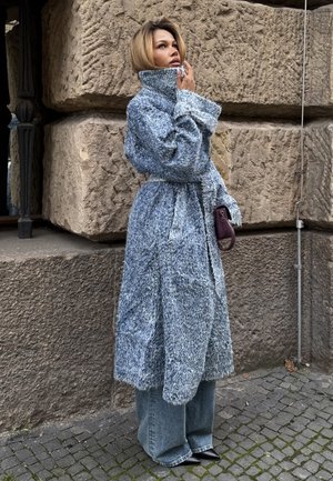 Übergroßer, strukturierter blauer Mantel mit hohem Kragen, tailliert und mit langen Ärmeln; kombiniert mit weitem Jeans und Schuhen mit Absatz; hält eine kleine Tasche.