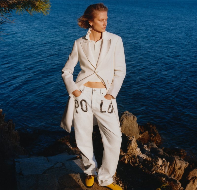 Mujer de pie en la costa rocosa, vestida con blazer blanca, pantalones blancos con el texto "POLO" y zapatos amarillos, con el océano de fondo.