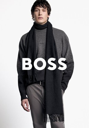 Schwarzer Schal mit Fransen, über einem dunkelgrauen Pullover geschichtet. Der Pullover hat ein dezentes Farbverlauf-Muster. Das Model steht vor einem weißen Hintergrund.