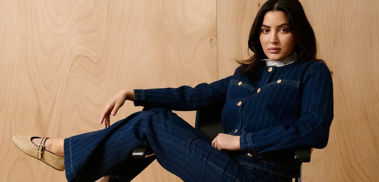 Jeune femme aux cheveux foncés portant un costume en denim bleu foncé et des ballerines beiges, assise de manière détendue sur une chaise noire devant un fond en panneaux de bois.