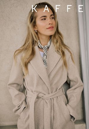 Frau mit langen blonden Haaren, die einen beigen Mantel mit Gürtel und einen gestreiften Halstuch trägt, steht vor einer hellen strukturierten Wand und schaut nach rechts.