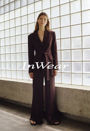 Frau in dunkelbraunem Blazer mit Gürtel und weitem Hosenbein steht barfuß vor einer Wand aus satiniertem Glasbaustein mit dem Schriftzug "InWear".