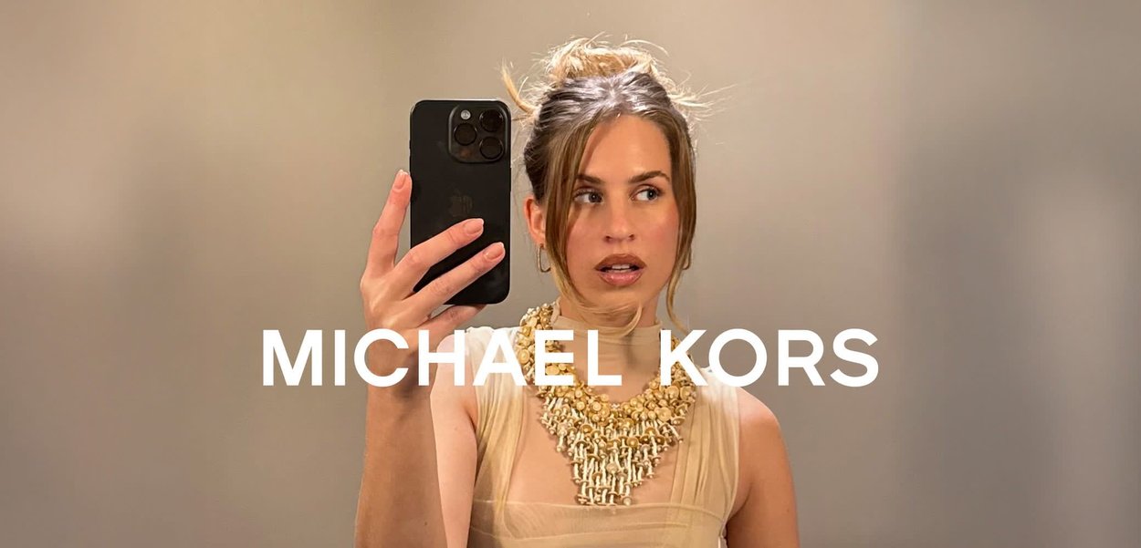 Mujer con peinado recogido, lleva un collar grueso de oro y una camiseta beige, sosteniendo un teléfono inteligente negro para un selfie, con el texto "MICHAEL KORS" superpuesto.
