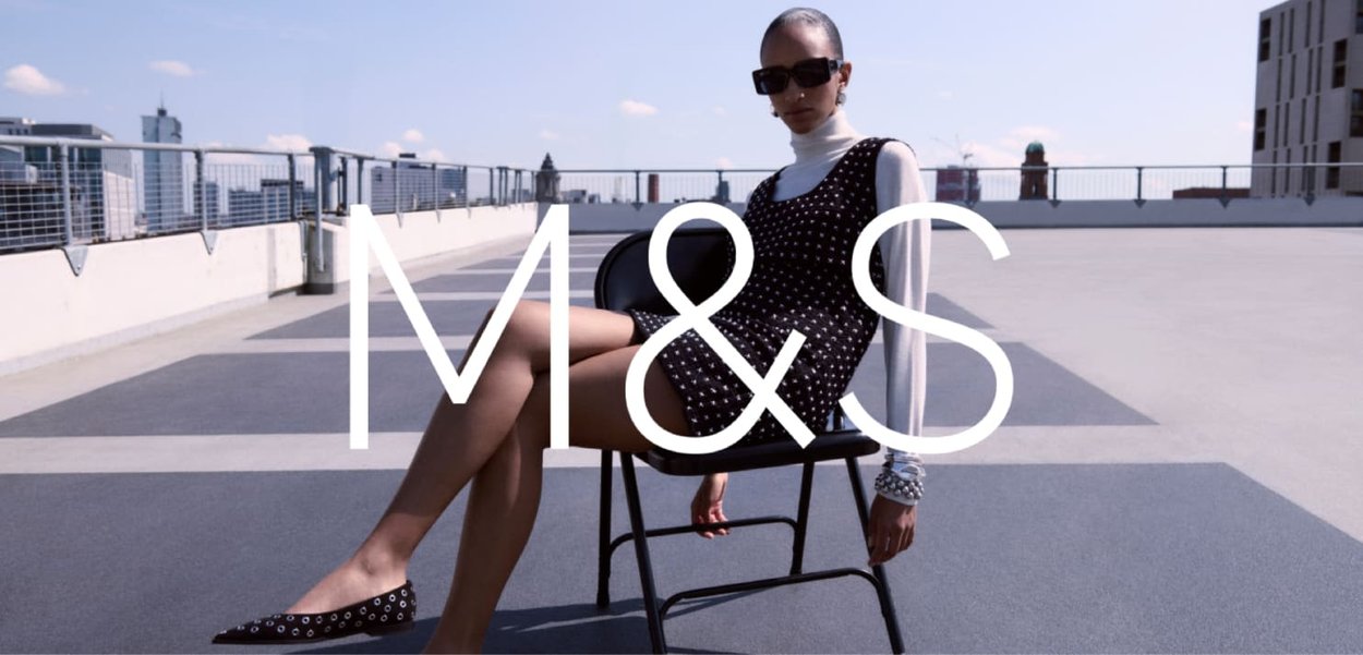Robe noire à pois blancs, de forme ajustée et décolletée en arrondi. Le modèle est assis sur une chaise noire, portant un col roulé blanc et des lunettes de soleil.