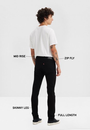 Hombre con tatuajes que lleva jeans ajustados negros de tiro medio y longitud completa, y camiseta blanca, de perfil con características de los jeans etiquetadas.