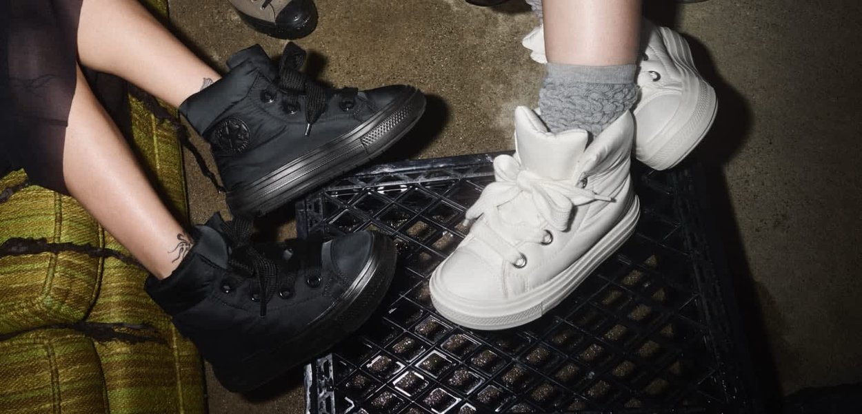 Due persone che indossano sneaker alte, una nera con caviglia tatuata e una bianca con calzini grigi, appoggiano i piedi su una cassa nera.