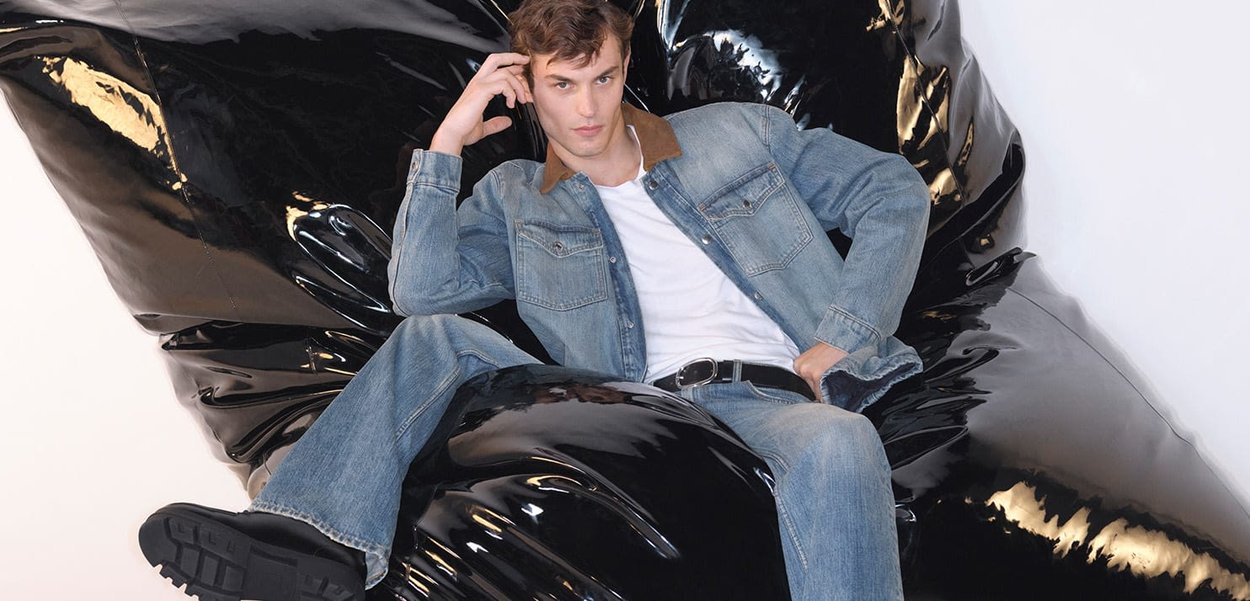 Jeansjacke mit braunem Kragen, weißem T-Shirt und blauen Jeans, getragen von einer Person, die auf einem großen schwarzen, glänzenden Sitzsack sitzt.