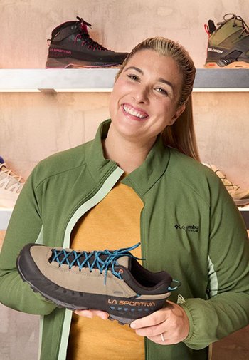 Braun und schwarze Wanderschuhe mit blauen Schnürsenkeln, strukturiertem Profil und "LA SPORTIVA" Branding. Getragen von einer lächelnden Person in einer grünen Jacke.
