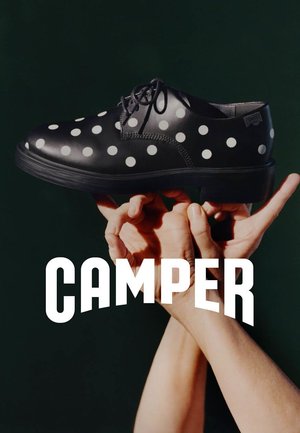Ręce trzymające czarne skórzane buty w białe kropki nad białym napisem "CAMPER" na ciemnozielonym tle.