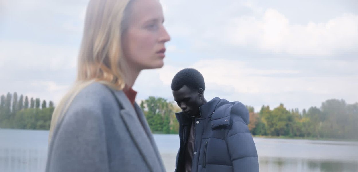 Due modelli vicino a un corso d'acqua. Uno indossa un blazer grigio chiaro, mentre l'altro indossa una giacca imbottita scura sopra una maglietta marrone.