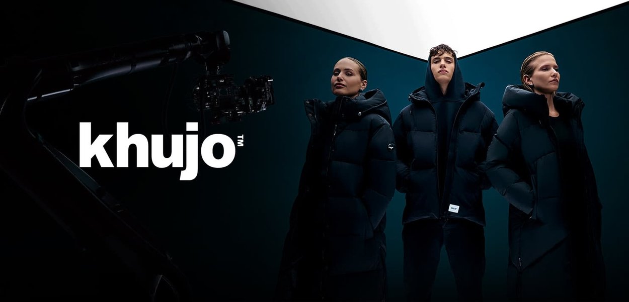 Drie modellen dragen zwarte pufferjassen met capuchons. De achtergrond is donker met een reflecterende lichtbron erboven, die de texturen van de jassen accentueert.