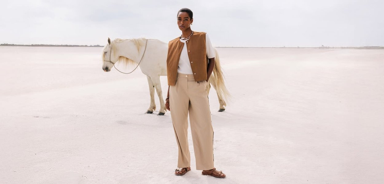 Tan mouwloze vest over een wit shirt, beige gestreepte wijde broek en bruine sandalen. Witte paard staat in een lichte, zandkleurige omgeving.