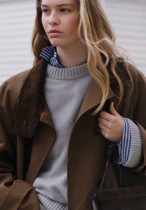 Młoda kobieta o długich blond włosach, ubrana w brązowy płaszcz na szarym swetrze i pasiastą koszulę, trzymająca ciemną torebkę, patrząca zamyślona w lewo.