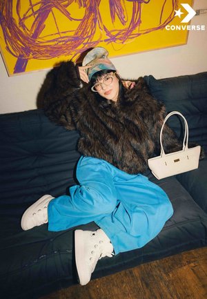 Chaqueta marrón de piel sintética, pantalones anchos azules, zapatillas blancas de caña alta y un bolso blanco. Posición relajada en un sofá oscuro con arte abstracto de fondo.