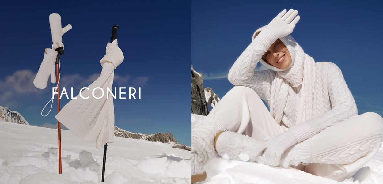 Muffole, sciarpa e cappello bianchi a maglia appoggiati a dei bastoncini da sci nella neve accanto a una persona vestita con abbigliamento bianco a maglia seduta in un paesaggio montano innevato.