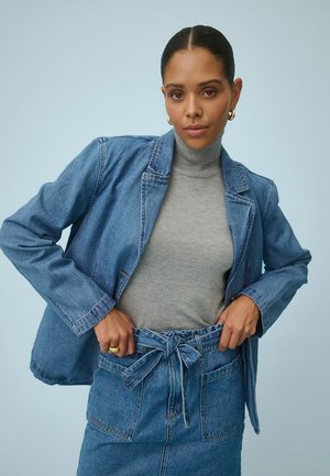 Frau trägt einen grauen Rollkragenpullover, ein Jeansblazer und einen hoch taillierten Jeansrock mit gebundenem Gürtel, posiert vor einem hellblauen Hintergrund.