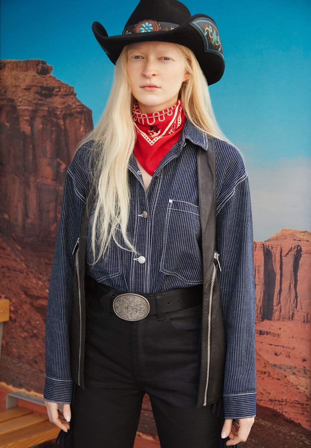 Rayado azul camisa de botones, chaleco de cuero negro, bandana roja, hebilla de cinturón plateada y pantalones negros. Presenta un sombrero de vaquero con acentos decorativos.