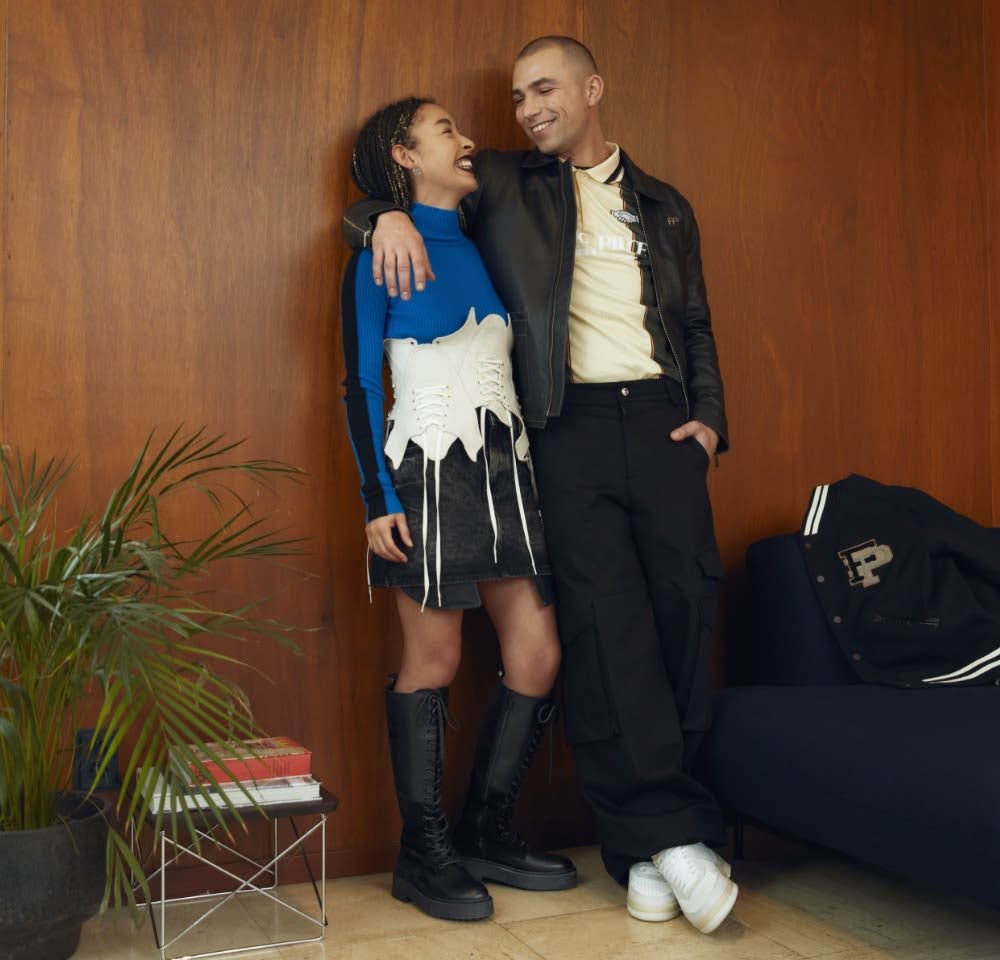 Twee personen staan samen in een moderne binnenomgeving. De vrouw draagt een blauwe blouse met lange mouwen, een gelaagde witte korset en een zwarte rok met kniehoge laarzen. De man heeft een zwart leren jack aan over een beige shirt en zwarte cargobroek met witte sneakers.