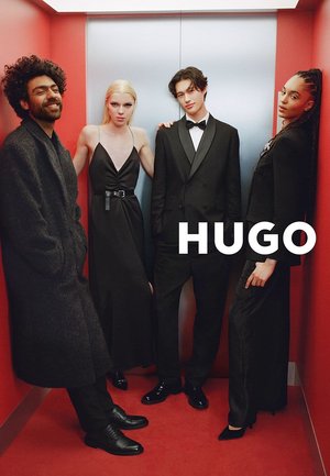 Quatre mannequins divers vêtus de tenues noires formelles posent dans un ascenseur aux murs rouges avec des portes argentées, avec le texte "HUGO" visible.