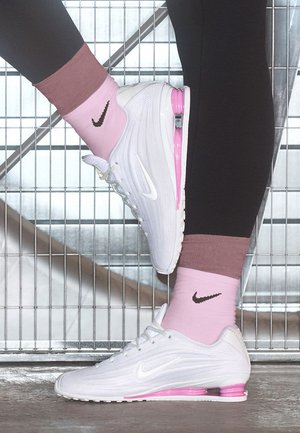 Gambe con leggings neri e calzini rosa Nike, indossando scarpe da ginnastica Nike bianche e rosa, un piede poggiato su una griglia metallica di sfondo.