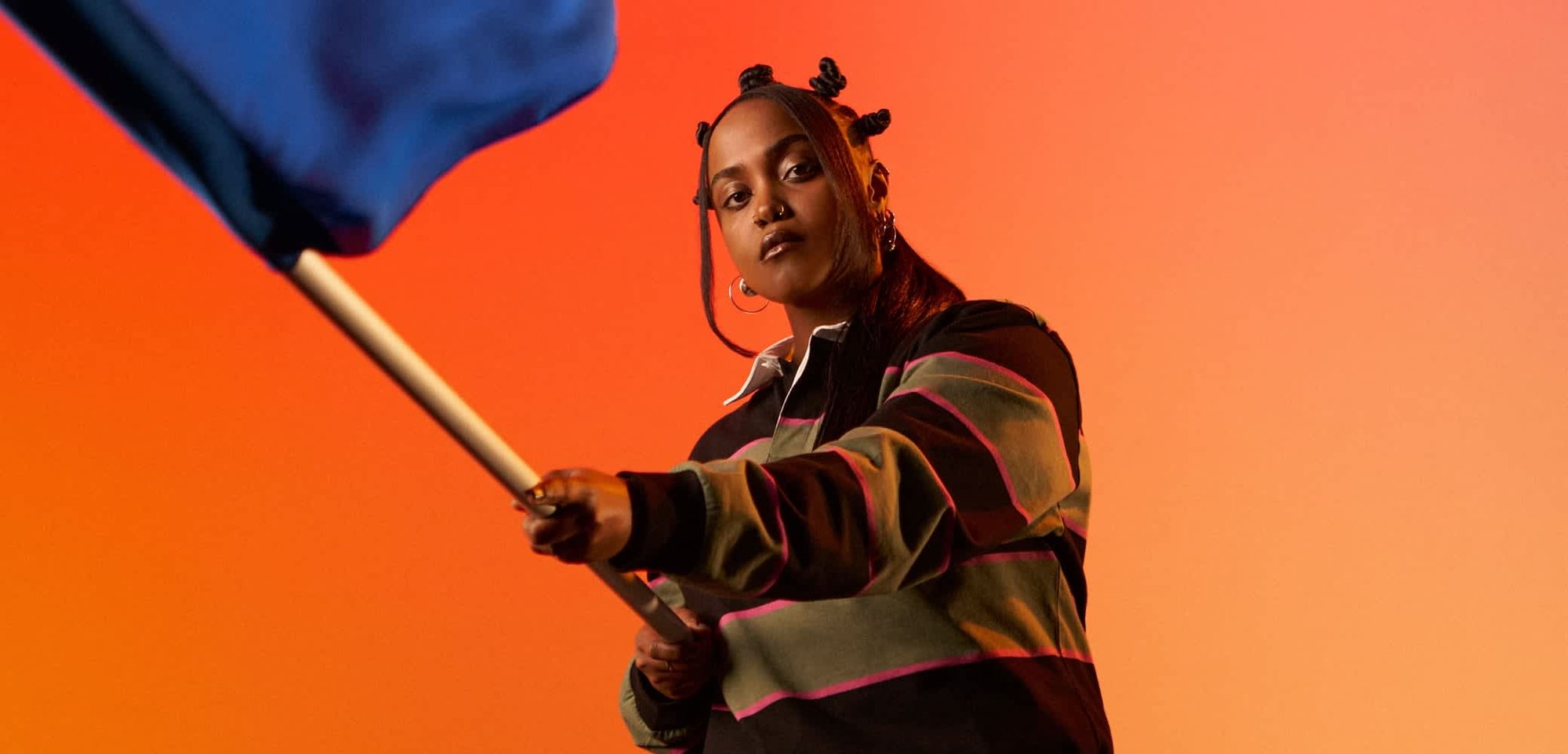 Model, die eine große blaue Flagge hält, trägt ein gestreiftes Sweatshirt in Grün und Schwarz mit pinken Akzenten sowie einen langen, cremefarbenen Rock. Der Hintergrund ist ein gradient orange.