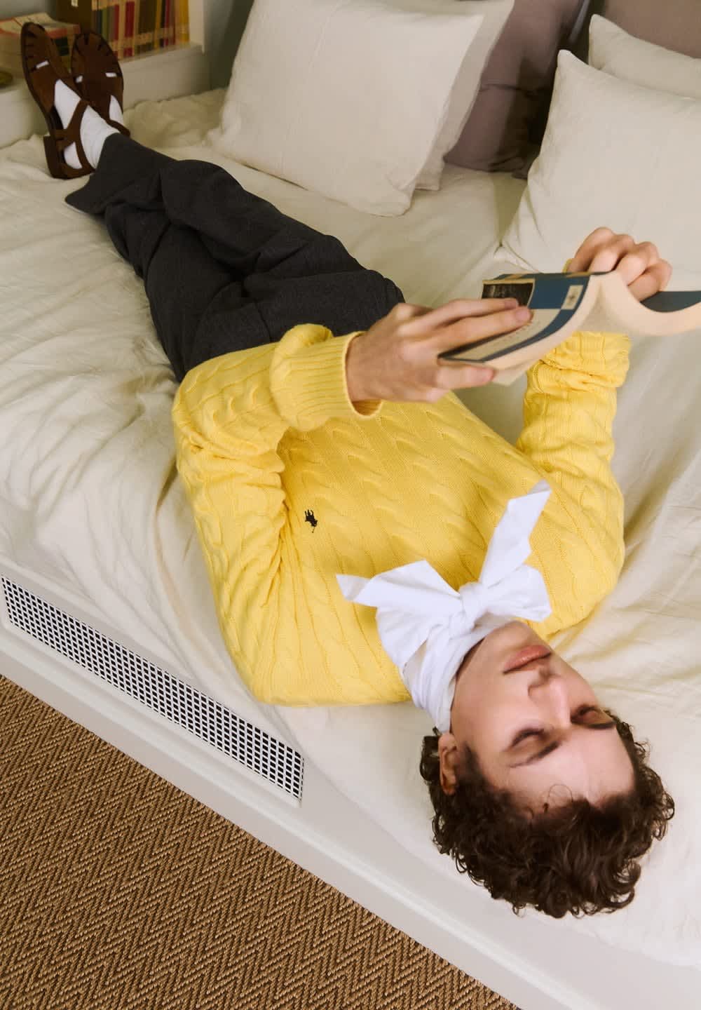 Person som ligger på en säng med en gul tröja, vit scarf, mörka byxor och sandaler med strumpor, och läser en bok medan han vilar huvudet på kudden.