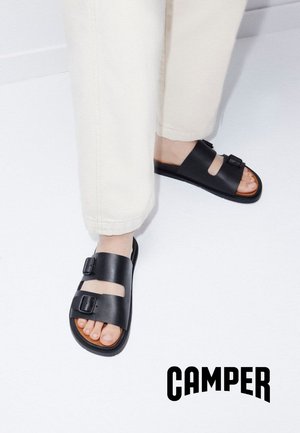 Pies con sandalias de cuero negras de doble hebilla combinadas con pantalones color crudo sobre un suelo blanco con el texto "CAMPER" debajo.