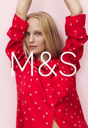 Blondynka w czerwonej koszuli w białe wzory serc, unosząca ręce nad głową na tle bladego różu.