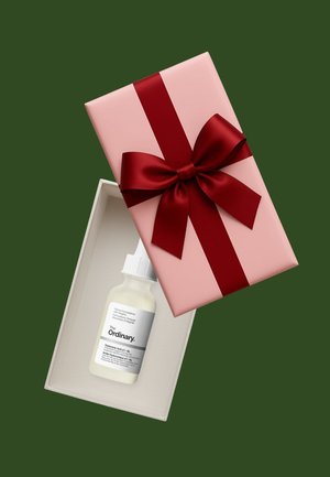 Piccola bottiglia bianca di siero per la cura della pelle The Ordinary all'interno di una scatola regalo rosa aperta con un grande fiocco rosso su sfondo verde.