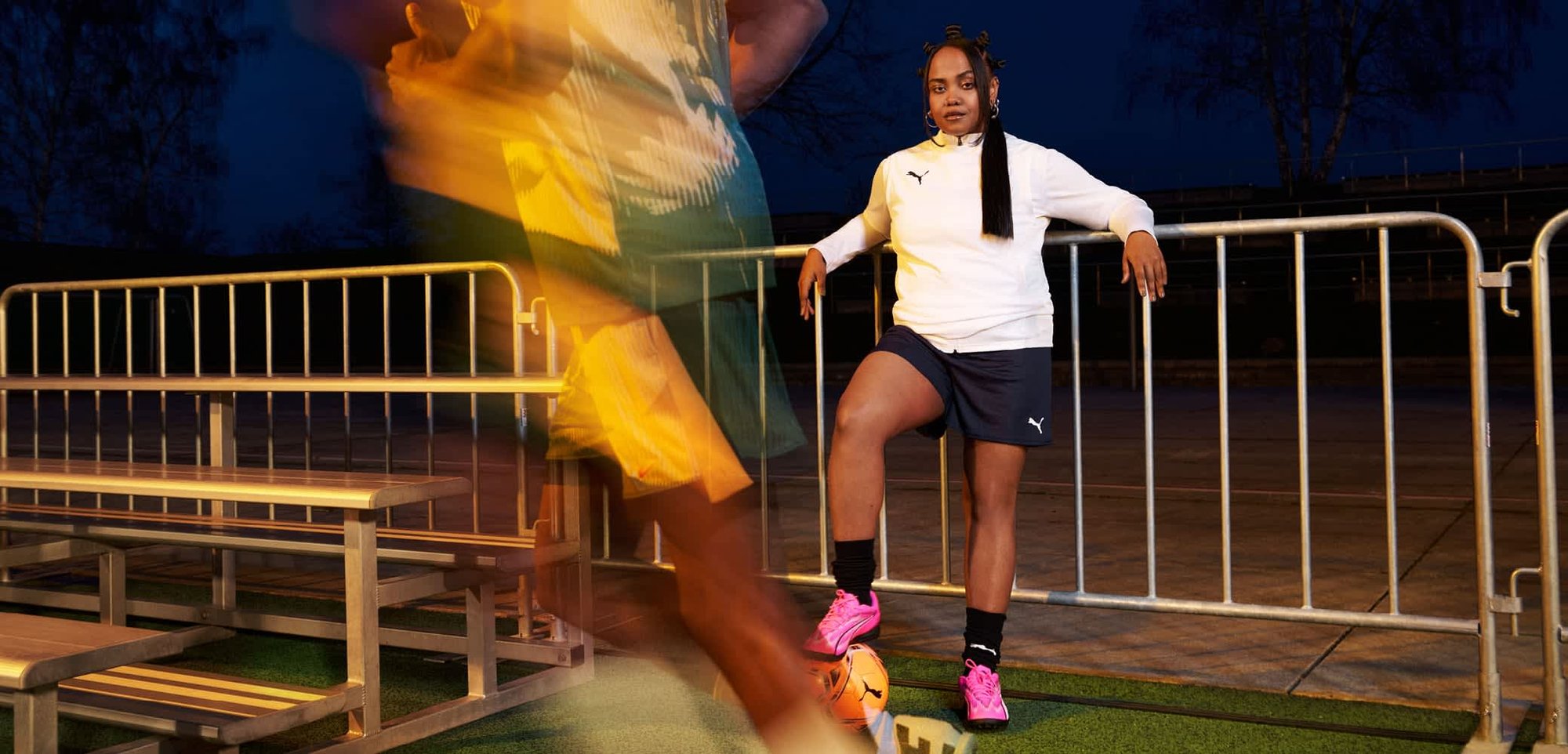 Zwei Personen in Sportkleidung auf einem Feld. Eine trägt einen weißen Sweatshirt und schwarze Shorts; die andere ein gelbes und grünes Trikot.