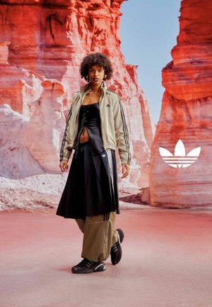 Femme aux cheveux bouclés portant une tenue de sport Adidas, debout sur un sol rose avec des formations rocheuses rouges et un ciel bleu clair en arrière-plan.