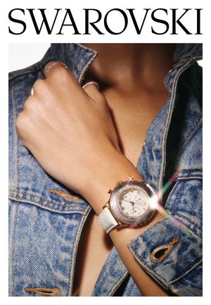 Montre Swarovski avec un bracelet blanc, un cadran rond, des accents dorés et une lunette en cristal, portée sur une main avec des manches de veste en denim visibles.