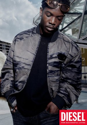 Giubbino bomber con colletto in tessuto grigio e nero a fantasia, indossato sopra una maglietta nera. Gli accessori includono occhiali da sole colorati sulla testa.