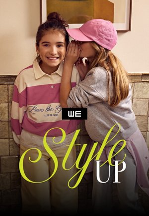 Twee meisjes binnen, één fluistert naar de ander terwijl ze glimlacht, gekleed in casual pastel kleding en een roze pet. Tekst 'WE Style UP' staat over de afbeelding.