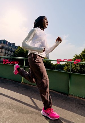 Athlet i hvid langærmet top og brune joggebukser, iført lyserøde Nike løbesko, jogger på en sti med grønt i baggrunden.