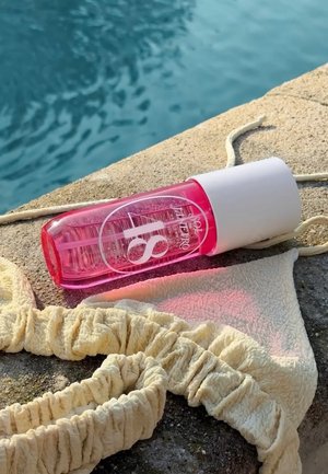 Pink sunscreen bottle and beige textured bikini top resting on a stone surface beside clear blue water.