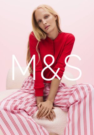 Femme aux longs cheveux blonds portant un cardigan rouge et un pantalon large rose avec des rayures rouges, assise sur une chaise rose clair devant un fond rose.